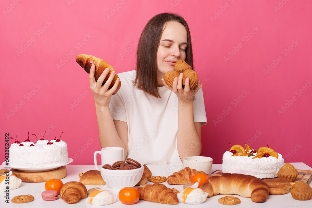 Enjoyable woman wearing white T-shirt sitting at festive table with homemade confectionery smelling tasty pastry isolated over pink background. Enjoyable woman wearing white T-shirt sitting at festive table with homemade confectionery smelling tasty pastry isolated over pink background.