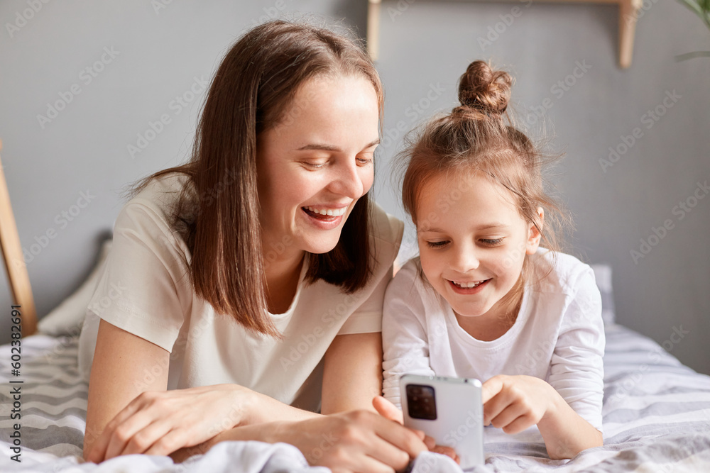 Portrait happy daughter and mother laying on bed playing smartphone watching video or photos in social networks expressing positive emotions. Portrait happy daughter and mother laying on bed playing smartphone watching video or photos in social networks expressing positive emotions.