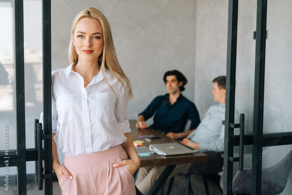 Portrait of attractive blonde female office worker in casual clothes standing in modern office room, confident looking at camera. Startup business team discussing project sitting at desk on background Portrait of attractive blonde female office worker in casual clothes standing in modern office room, confident looking at camera. Startup business team discussing project sitting at desk on background