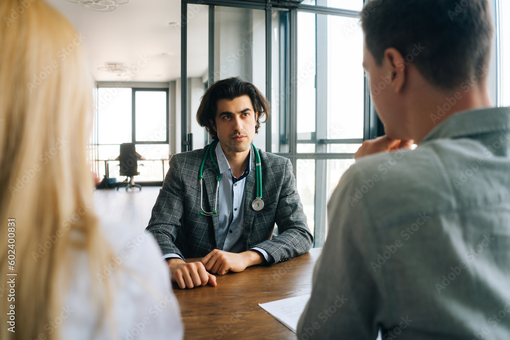 Obraz premium Portrait of professional male doctor with stethoscope talking to unrecognizable man and woman about health during counseling at medical office. Serious physician offering advices to young couple.