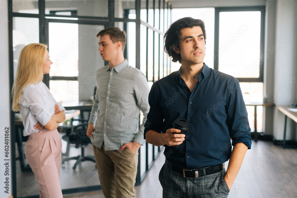 Portrait of successful startup businessman in casual clothes holding takeaway coffee cup in hand standing in office, confident looking away. Young business team friendly chatting on background Portrait of successful startup businessman in casual clothes holding takeaway coffee cup in hand standing in office, confident looking away. Young business team friendly chatting on background