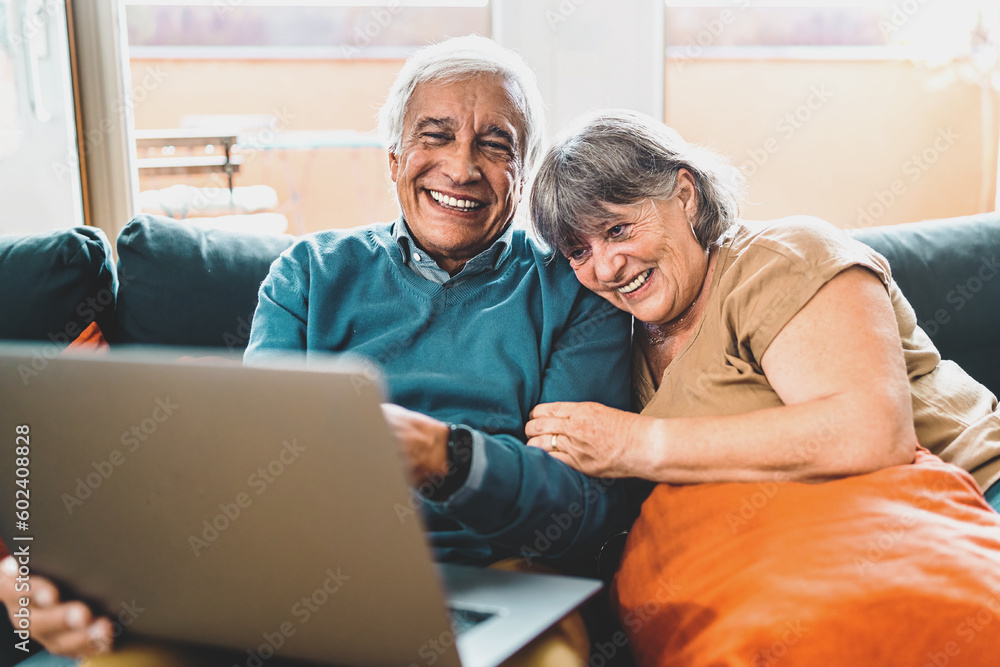 Happy Retired Couple Watching Content on Laptop - A joyful couple sits on the couch, with the man holding a laptop on his lap as they embrace, laughing heartily at online content.