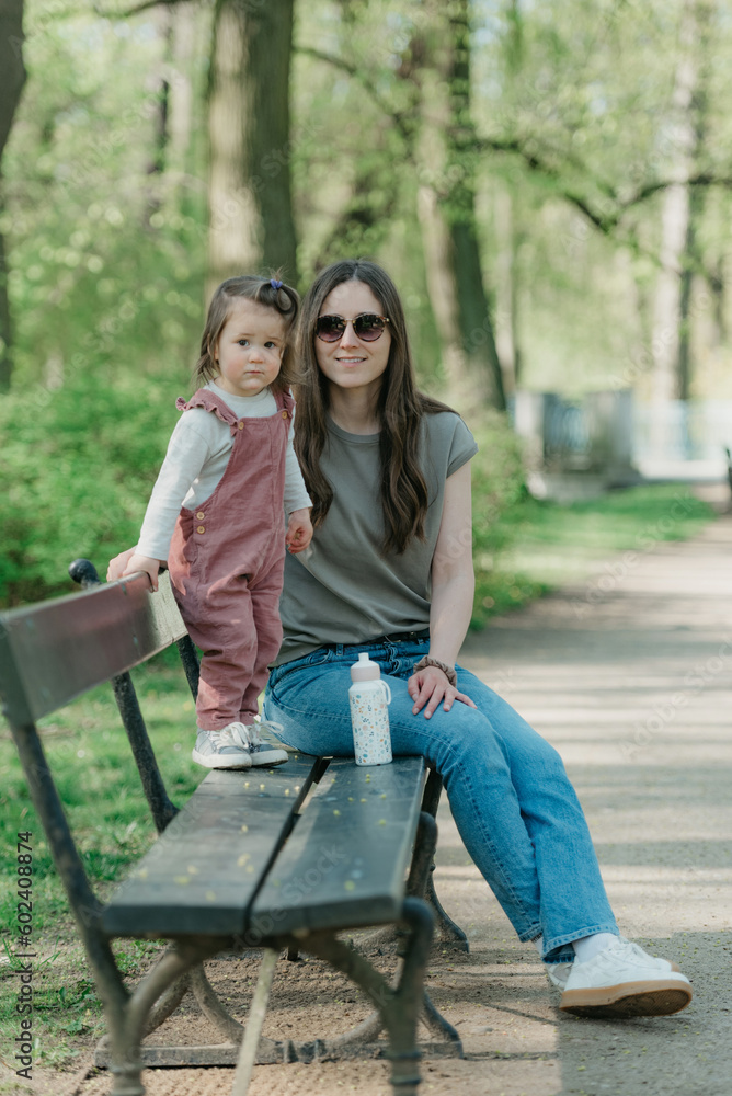 A female toddler in velvet overall is on the bench with her mother.