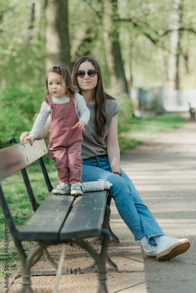 A female toddler in velvet overall is on the bench with her mother.