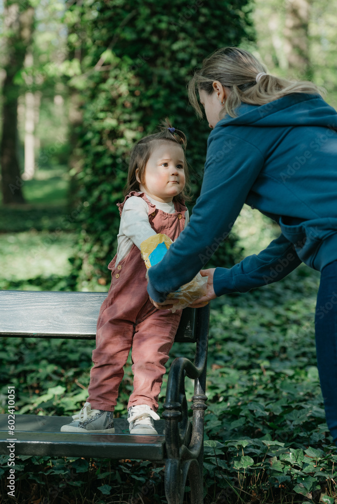 A female toddler in velvet overall is on the bench with her mother.