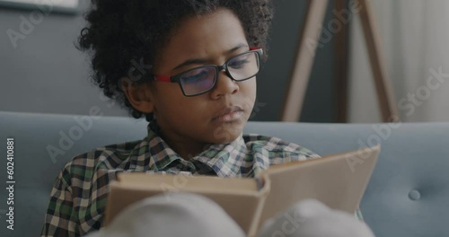 Slow motion of smart African American boy in eyeglasses reading book and smiling at home. Literature and education for children concept.