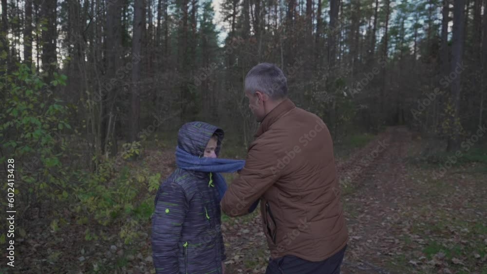 Father puts scarf on top of jacket for son among trees in autumn forest because of cold weather. Dad takes care of son and worries about health Father puts scarf on top of jacket for son among trees in autumn forest because of cold weather. Dad takes care of son and worries about health
