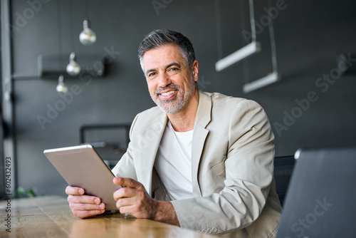 Smiling mid aged successful business man using digital tablet computer sitting in modern office. Happy mature older professional businessman entrepreneur working on technology device, portrait.