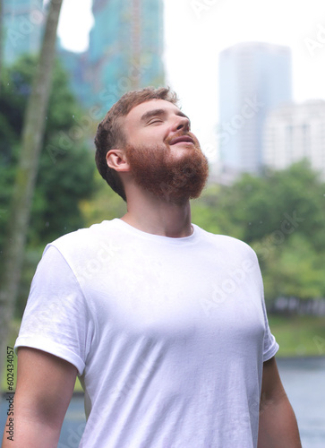Positive young man smiling during the rain in the park. Cheerful male enjoying the rain outdoors. Guy looking up and catching the rain drop with hands. Breath deep and relax