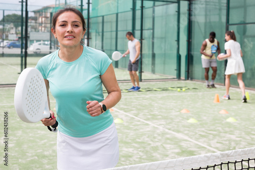 Happy woman learning to play padel game on tennis court outdoor. Other athletes are training in the background