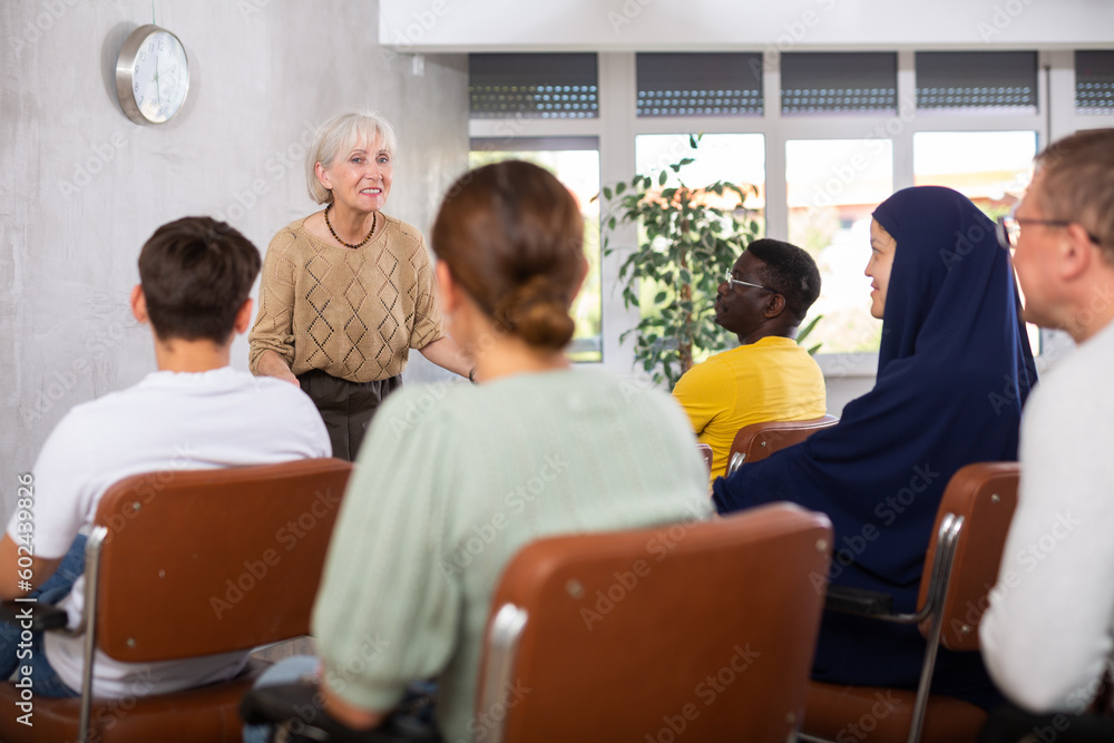 Fototapeta premium Emotional aged female tutor leading educational class for group of men and women of different nationalities sitting in auditorium