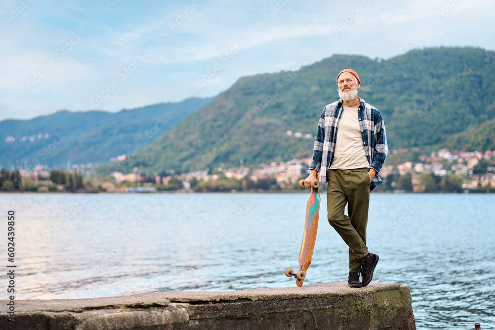 Active cool bearded old hipster man standing in nature park holding skateboard. Mature traveler skater enjoying freedom spirit and extreme sports hobby leisure lifestyle on lake background.