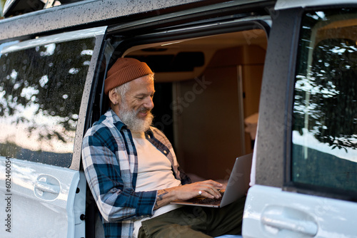 Happy older man sitting in rv camper van using laptop. Smiling mature active traveler holding computer on lap remote working online and enjoying vanlife, freedom, resting in outdoor camping.
