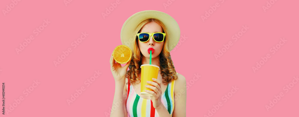 Summer portrait of stylish young woman drinking fresh juice with slice of orange fruits wearing straw hat, sunglasses on pink background
