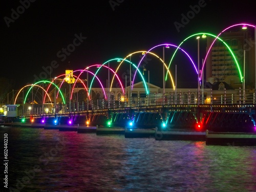 The Dutch-style buildings lining Handelskade, a street along St Anna Bay, as the Queen Emma Bridge glows in rainbow colors over the waters.