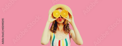 Summer portrait of cheerful young woman covering her eyes with fresh slices of orange fruits and looking for something wearing straw hat on pink background