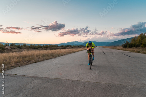  Triathlete riding his bicycle during sunset  preparing for a marathon. The warm colors of the sky provide a beautiful backdrop for his determined and focused effort.