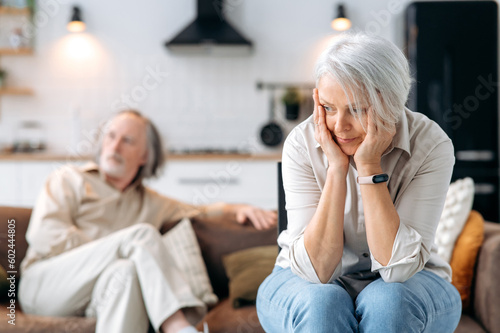 Slika na platnu Mature caucasian spouses misunderstanding each other, experiencing crisis in relationship, offended wife looks away disappointedly, the husband sits in defocus on background