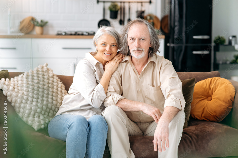 Happy together. Modern pensioners. Middle aged caucasian married couple, grey haired spouses spend time together in living room, wife and husband sit on a sofa, hugging, looking at camera, smile
