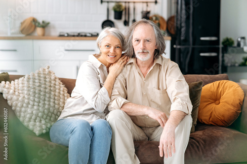 Happy together. Modern pensioners. Middle aged caucasian married couple, grey haired spouses spend time together in living room, wife and husband sit on a sofa, hugging, looking at camera, smile