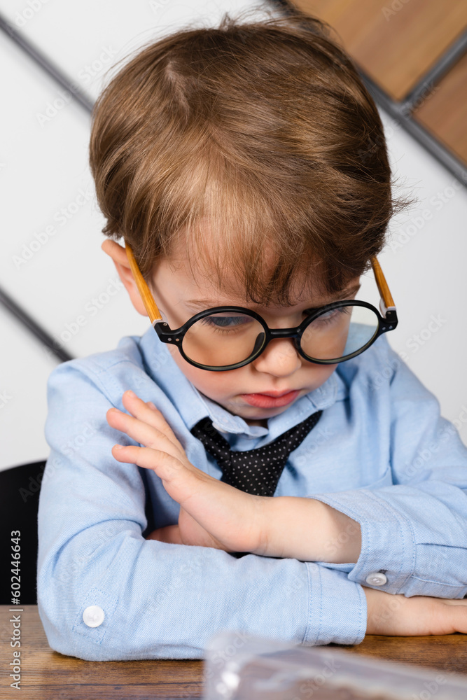 Sad Little boy in round eyeglasses, blue shirt and tie sitting at the desk. offended child sadly lowered their head. Sad Little boy in round eyeglasses, blue shirt and tie sitting at the desk. offended child sadly lowered their head.