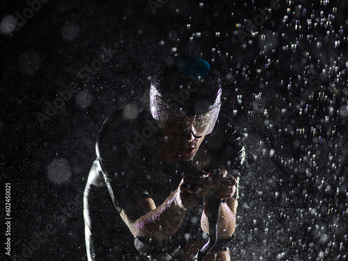A triathlete braving the rain as he cycles through the night  preparing himself for the upcoming marathon. The blurred raindrops in the foreground and the dark  moody atmosphere in the background add
