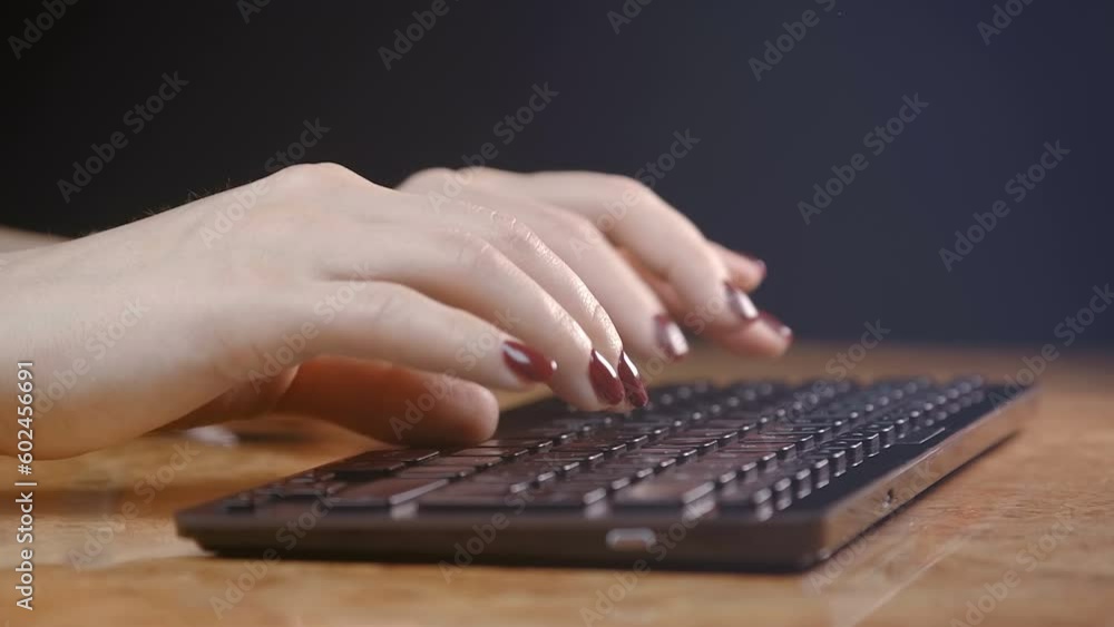 The hands of a young girl with beautiful shiny red nails are confidently typing on a black keyboard lying on a table with a wooden glossy top on a black background. Manicure with gel polish. Closeup The hands of a young girl with beautiful shiny red nails are confidently typing on a black keyboard lying on a table with a wooden glossy top on a black background. Manicure with gel polish. Closeup