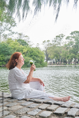 Mature Hispanic woman sitting in a park smiling while drinking green juice from a glass bottle. Concepts: healthy nourishment and lifestyle. © Carolina Jaramillo