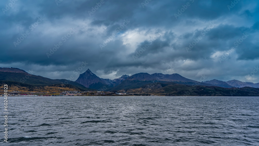 Picturesque Martial mountain range against a cloudy sky. City houses are visible on the shore. In the foreground is the water of the Beagle Canal. Argentina. Ushuaia. Tierra del Fuego Archipelago.