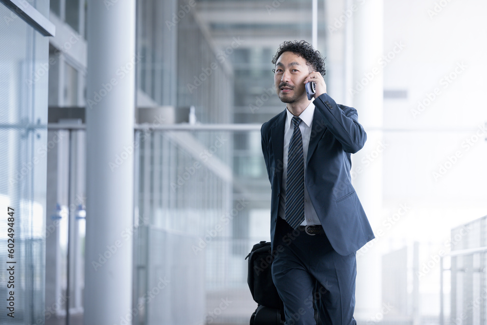 Middle-aged Asian male businessman in his 40s in a suit walking while pulling a carry-on case (suitcase) at an airport or train station. Middle-aged Asian male businessman in his 40s in a suit walking while pulling a carry-on case (suitcase) at an airport or train station.