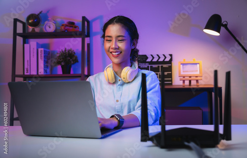 Fotografia Happy young Asian woman using laptop social media messages on the table for modem to high speed internet via LAN cable in modern home office at night