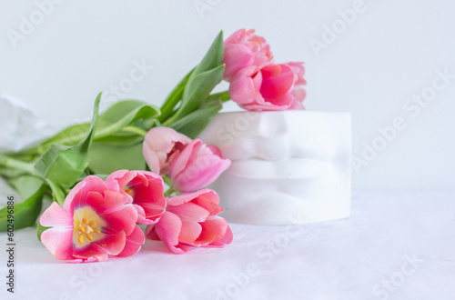 Pink tulips bouquet and plaster statue of lips lying on white wrinkled blanket background copy space