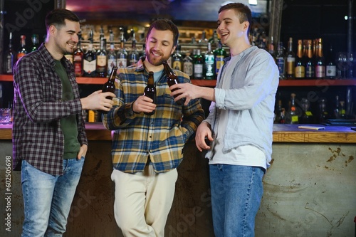 Three young men in casual clothes are smiling, holding bottles of beer while standing near bar counter in pub © Serhii