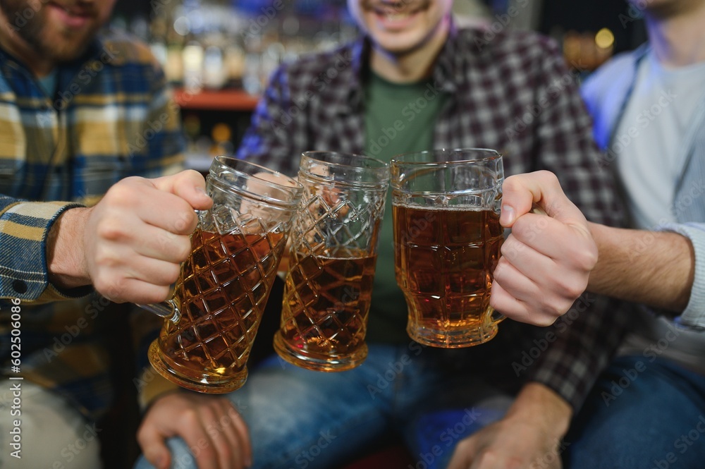 Three young men in casual clothes are smiling and clanging glasses of beer together while sitting at bar counter in pub