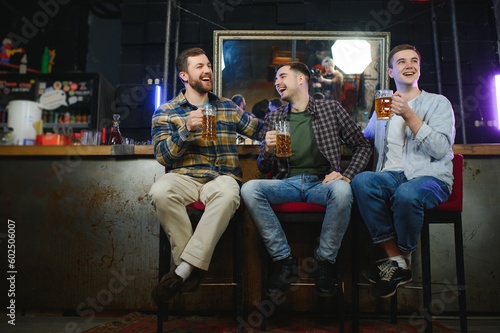 Three young men in casual clothes are smiling  holding bottles of beer while standing near bar counter in pub