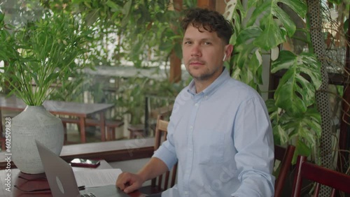 DescriptionMedium portrait shot of young businessman in wireless earphones and formal shirt sitting with laptop at table in outdoor cafe and posing on camera photo