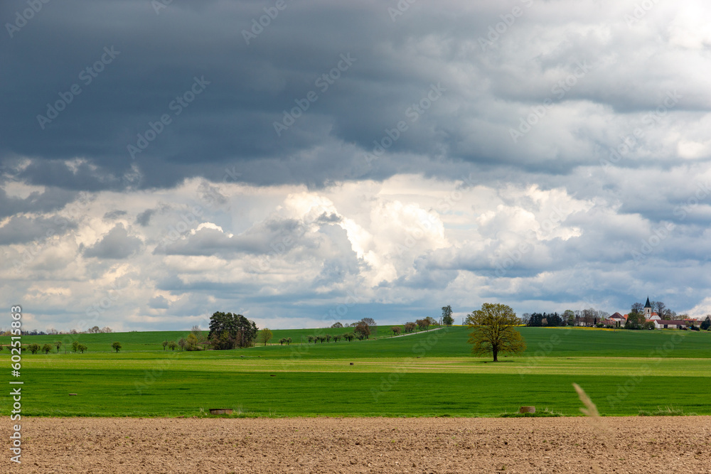 A small European village with a church on a hill, among spring fields, under a dramatic cloudy sky.