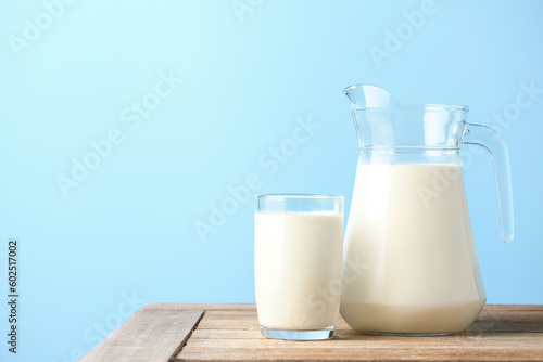 Glass and pitcher of fresh milk on wooden table with light blue background.