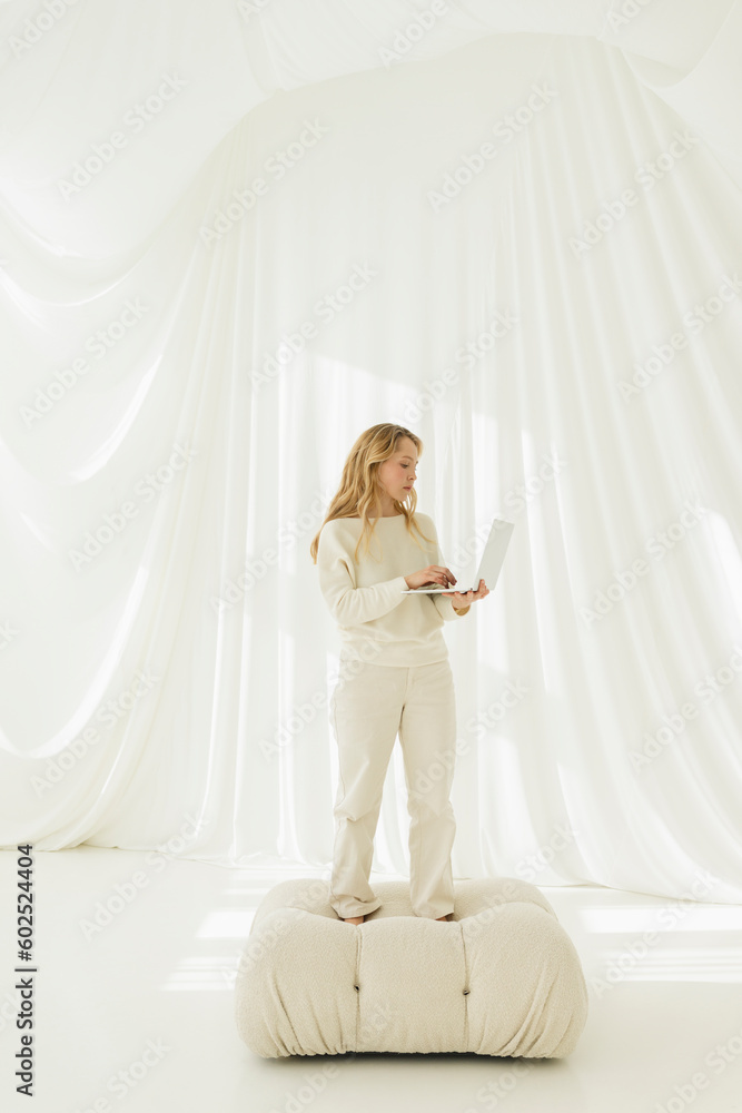 Blond woman using laptop standing on couch in front of translucent curtain Blond woman using laptop standing on couch in front of translucent curtain