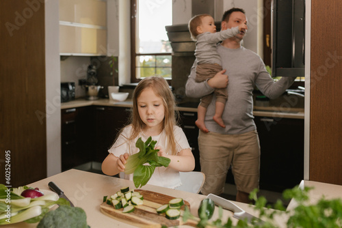 Girl holding Bok Choy with father carrying son in background at home photo