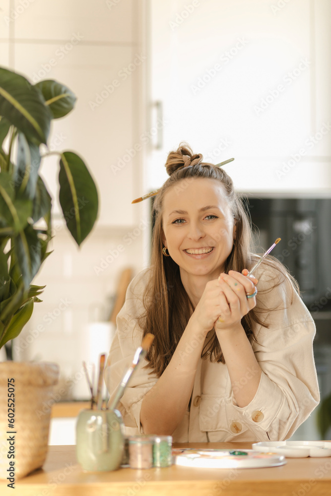 Smiling young woman holding paintbrush leaning on table at home Smiling young woman holding paintbrush leaning on table at home
