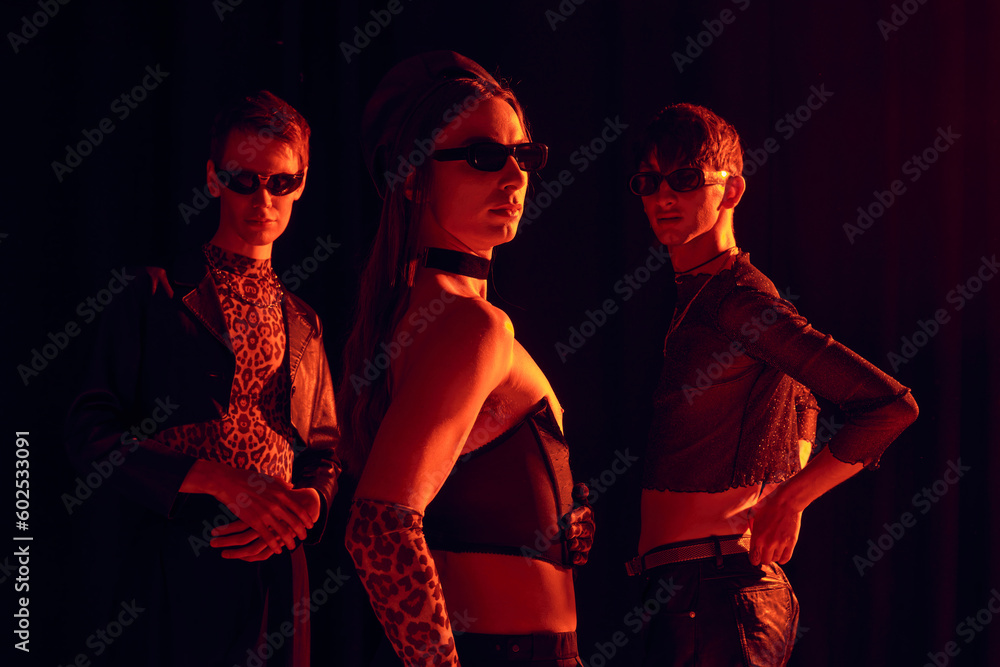 Group of fashionable homosexual community in sunglasses and party outfits posing together while celebrating lgbt month isolated on black with red lighting