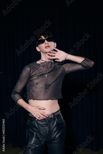 Fashionable homosexual man in sunglasses, sparkling top and necklaces posing during lgbt community month party isolated on black