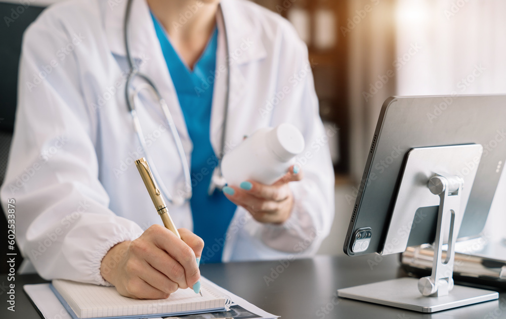 Fototapeta premium Pharmacist writing making notes in drug information using laptop computer sitting at desk. Woman physician, nurse or pharmacist wearing white coat writing in paper notebook.