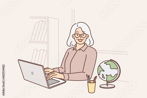 Elderly woman teacher uses laptop sitting at table with globe and teaching students via internet