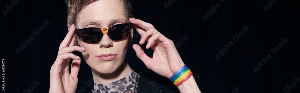 Portrait of trendy nonbinary person with lgbt flag on bracelet touching sunglasses during pride community month celebration isolated on black, banner Portrait of trendy nonbinary person with lgbt flag on bracelet touching sunglasses during pride community month celebration isolated on black, banner