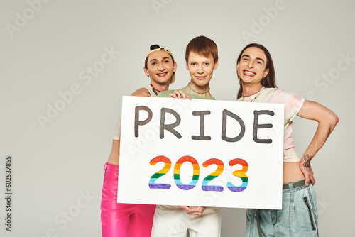 diverse and young lgbtq community friends with tattoos standing in colorful clothes and holding pride 2023 placard while looking at camera in studio, grey background