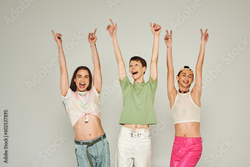 positive and young lgbt community friends in colorful clothes smiling while looking at camera, raising hands and celebrating pride month together on grey background in studio