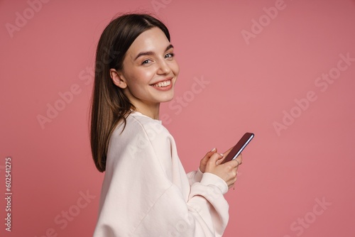 Woman using mobile phone and smiling at camera while standing isolated over pink background
