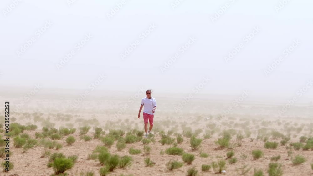 Young hipster man in sunglasses goes through semi-desert with rare plants during sandy storm. Slow motion Young hipster man in sunglasses goes through semi-desert with rare plants during sandy storm. Slow motion